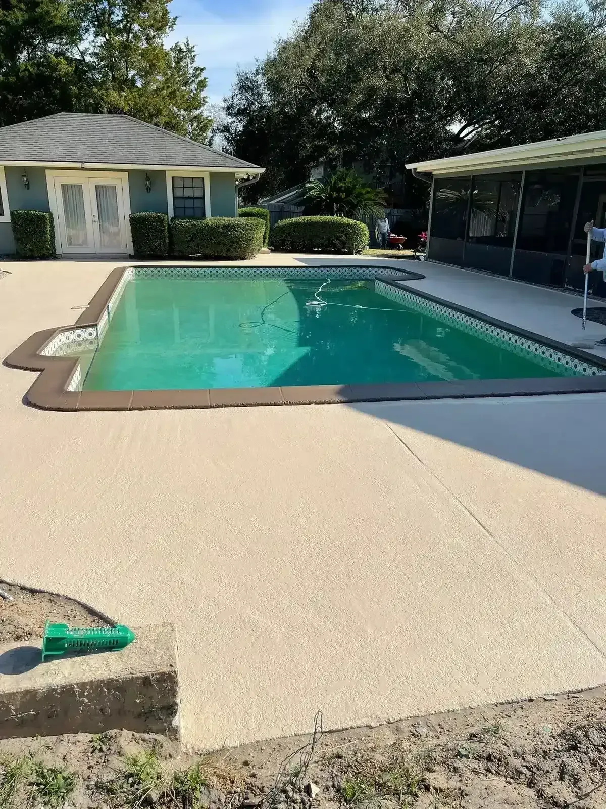 Backyard swimming pool with beige deck, white pool house, and covered patio in sunlight