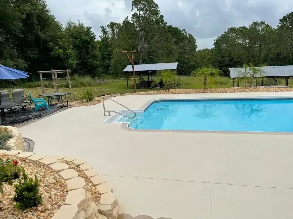 Outdoor swimming pool with blue water beside a beige patio and trees under a cloudy sky