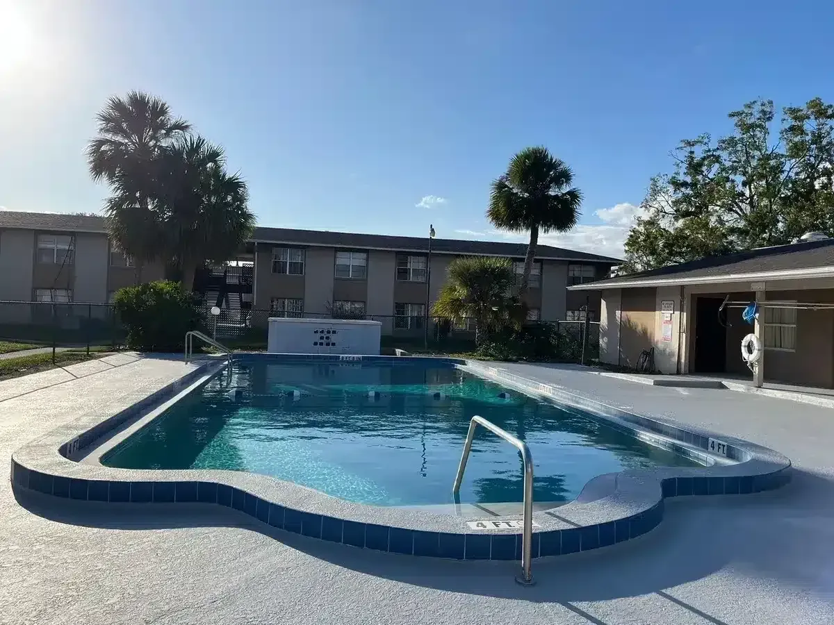 Apartment pool with clear blue water, white deck, and surrounding low-rise buildings under a sunny sky