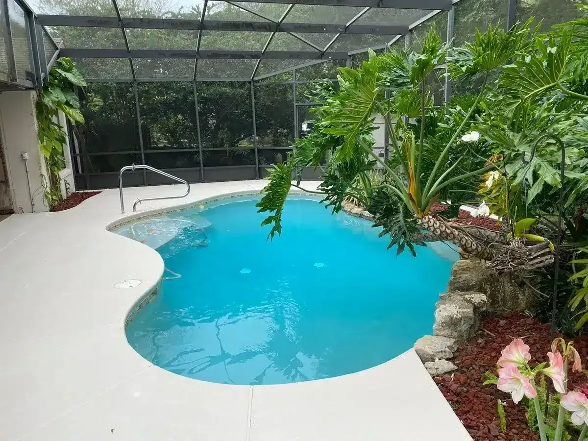 Screened pool with bright blue water, white deck, and lush tropical plants in a garden setting