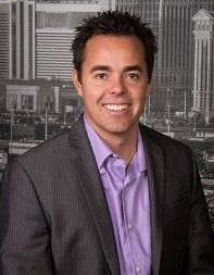 A man in a suit and purple shirt is smiling in front of a city skyline.