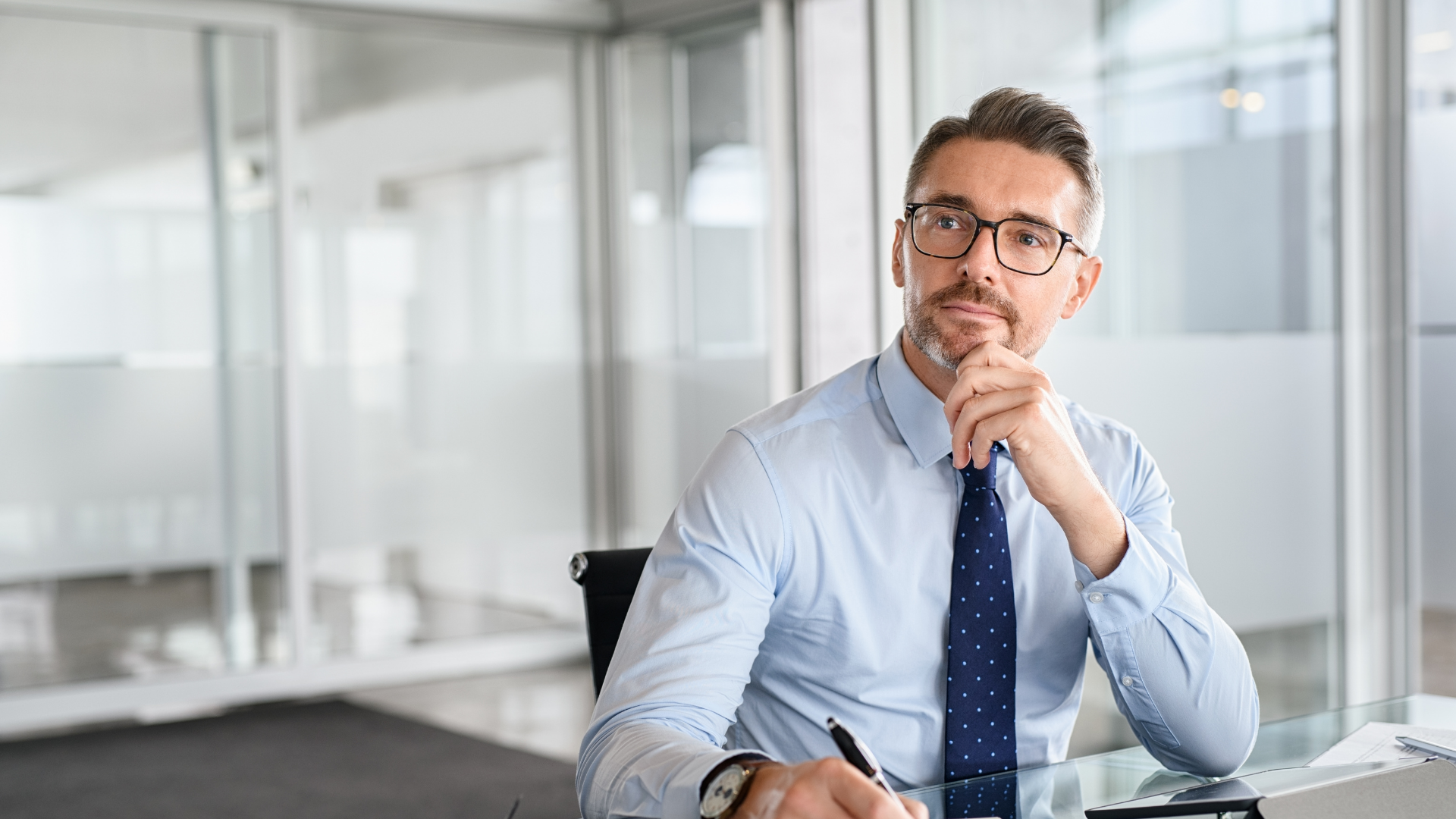 Man in blue shirt and tie, sitting and thinking, holding pen, in office.