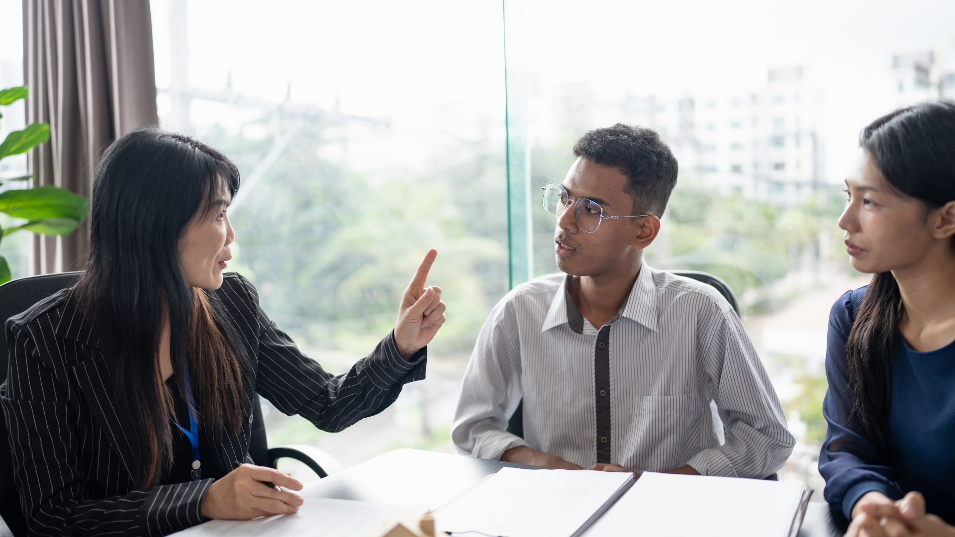 Woman pointing, addressing two people seated at a table; they appear engaged in a discussion.