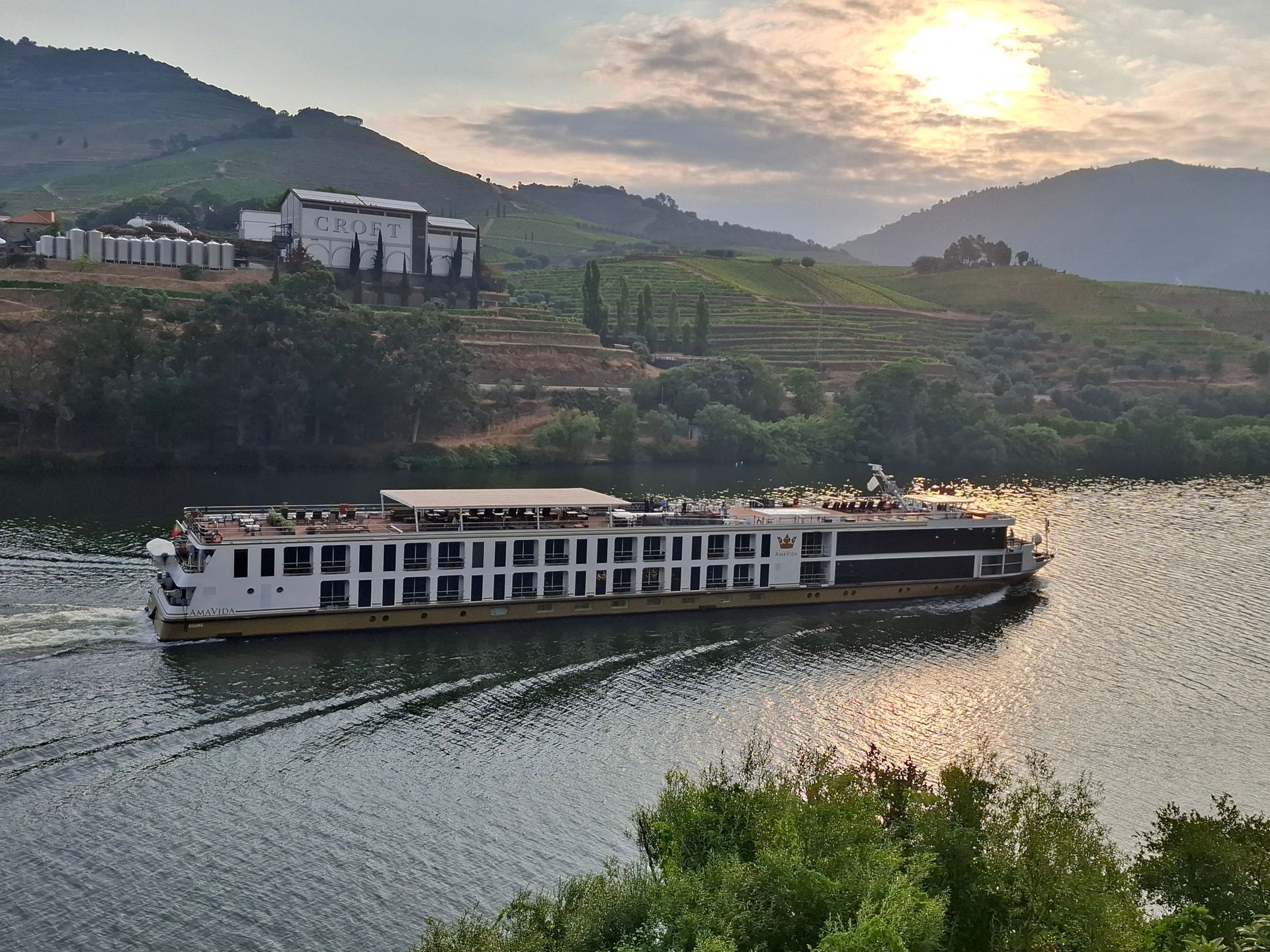 Bateau de passagers sur un lac au coucher du soleil, avec des collines et des vignobles en arrière-plan.
