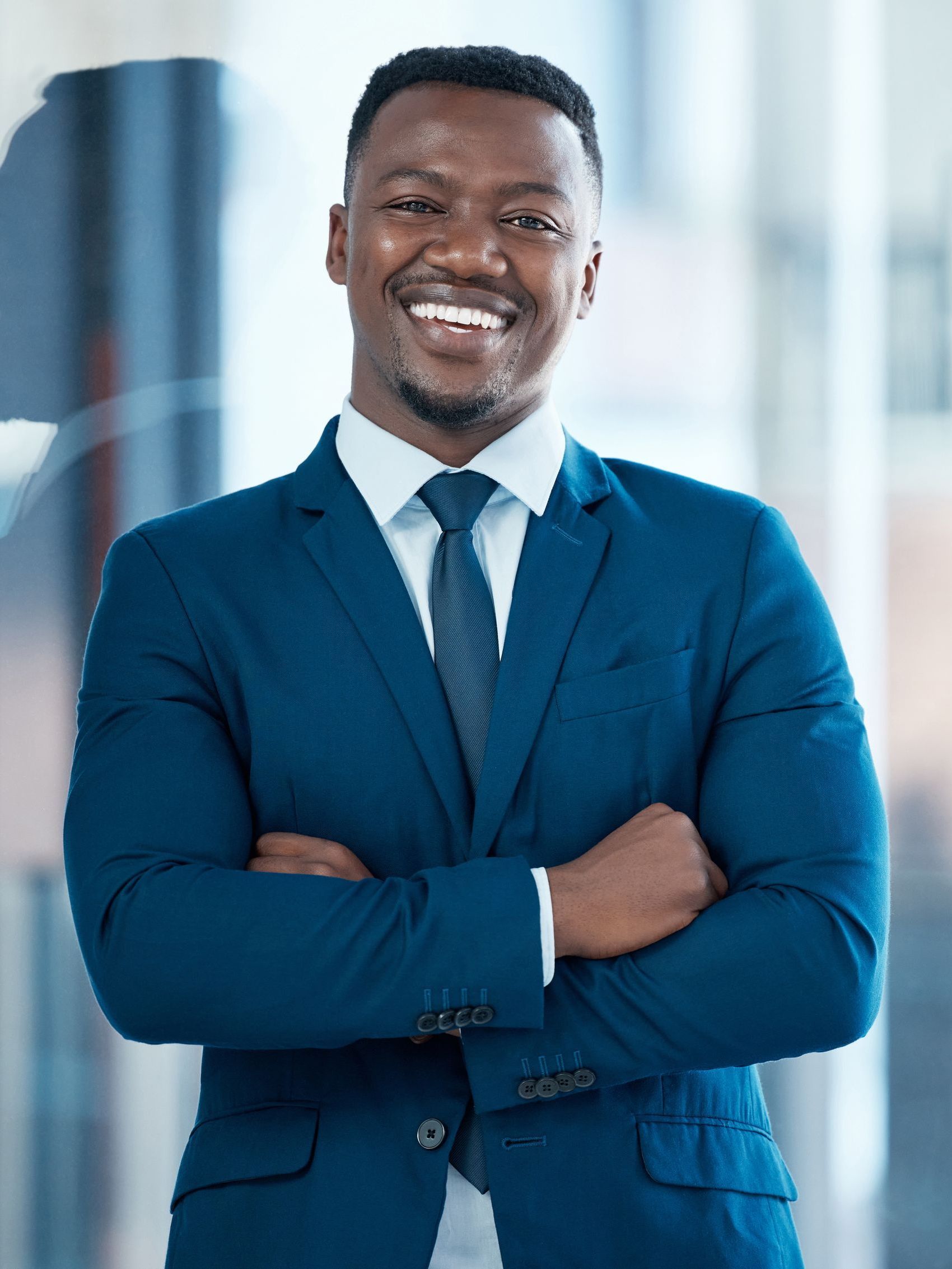 A man in a blue suit and tie is smiling with his arms crossed.