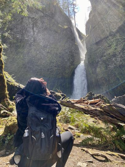 test Person with backpack kneels before waterfall in lush, sunlit canyon.