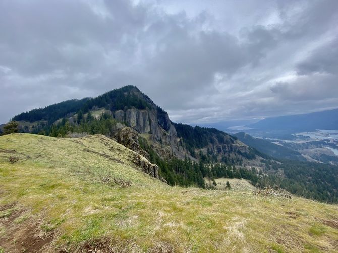 test Mountain summit under a cloudy sky. Cliffs and evergreen trees on the slope with a distant valley.
