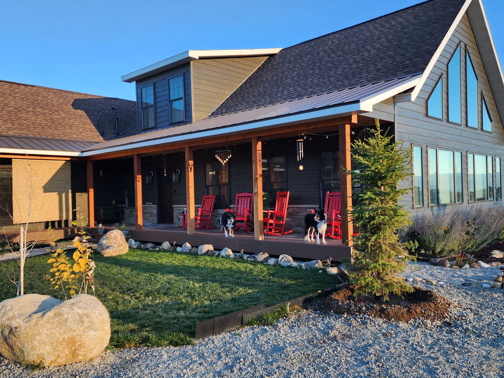 Modern cabin-style house with a covered porch, red chairs, and landscaped front yard at sunset