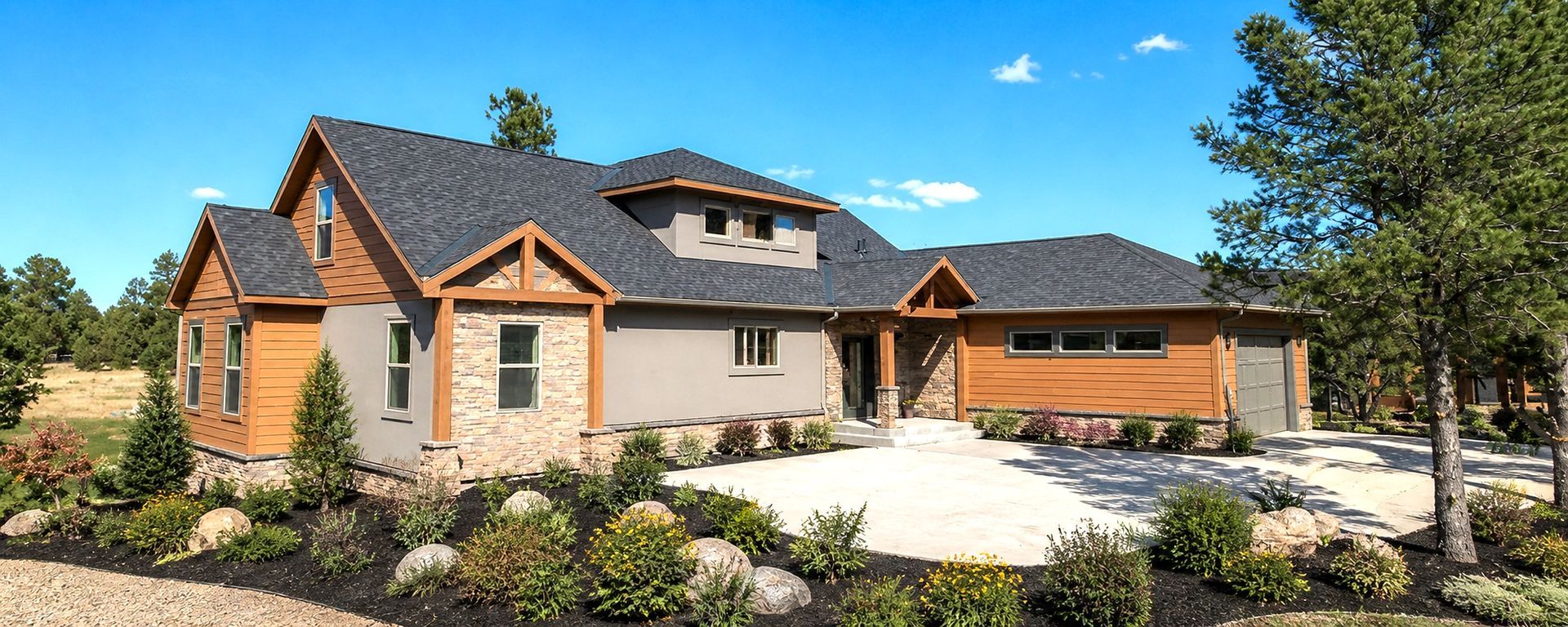 Modern stone-and-wood house with a large driveway, landscaped yard, and blue sky
