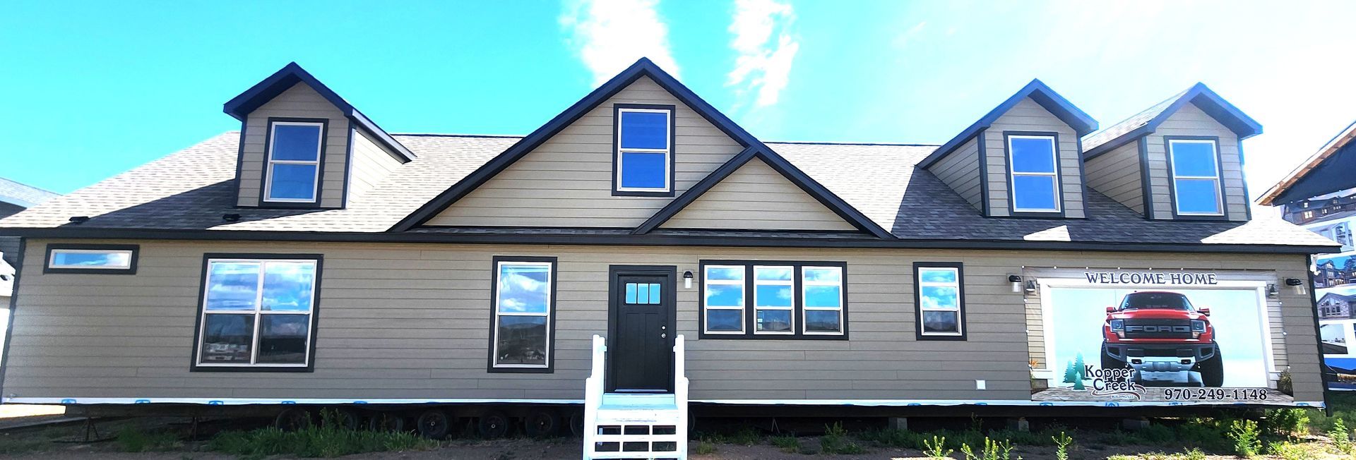 Wide tan house with dark roof, dormer windows, and a central white entrance under a blue sky.