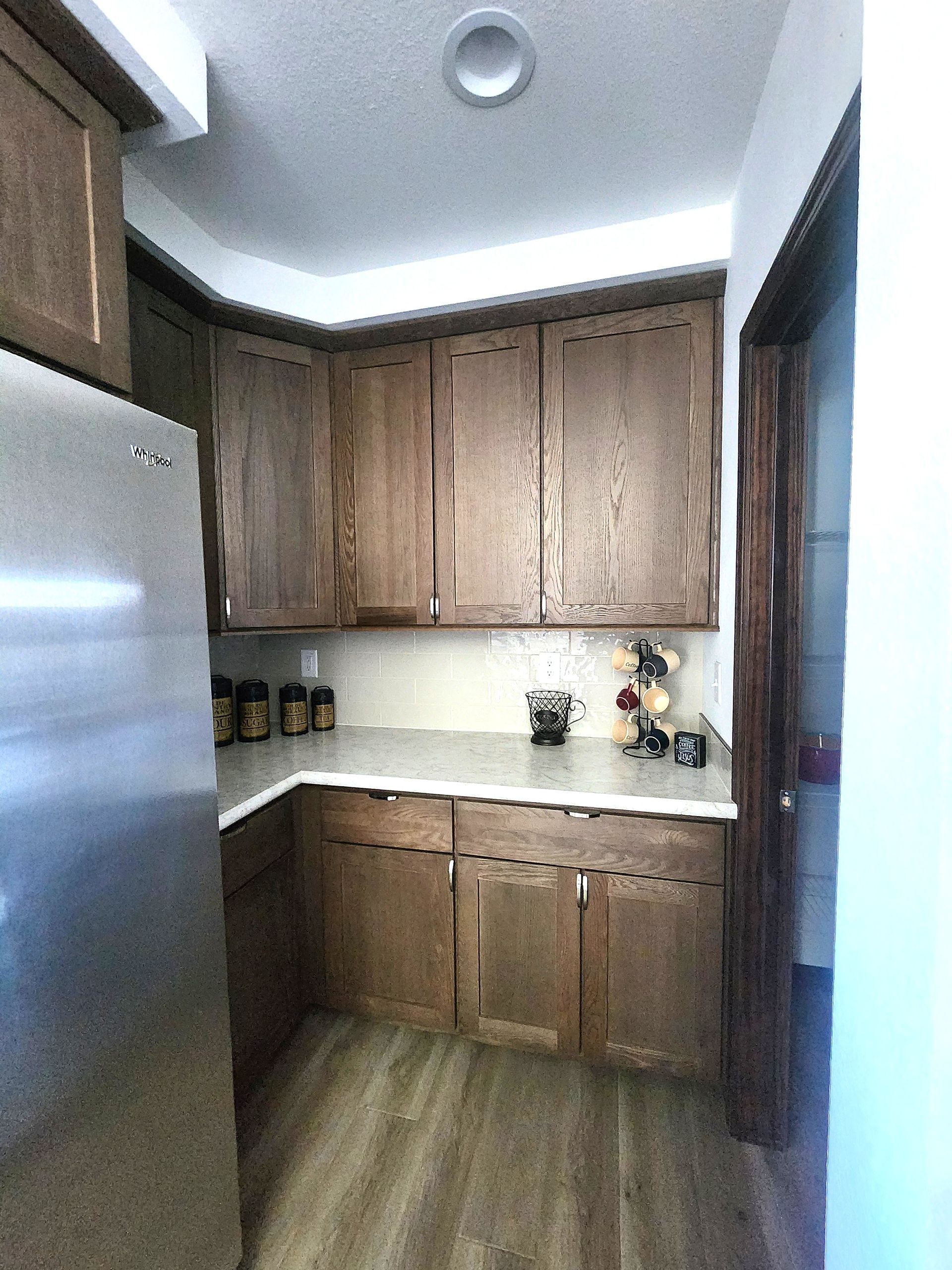 Small kitchen with wood cabinets, stainless refrigerator, white counters, and wood-look flooring.