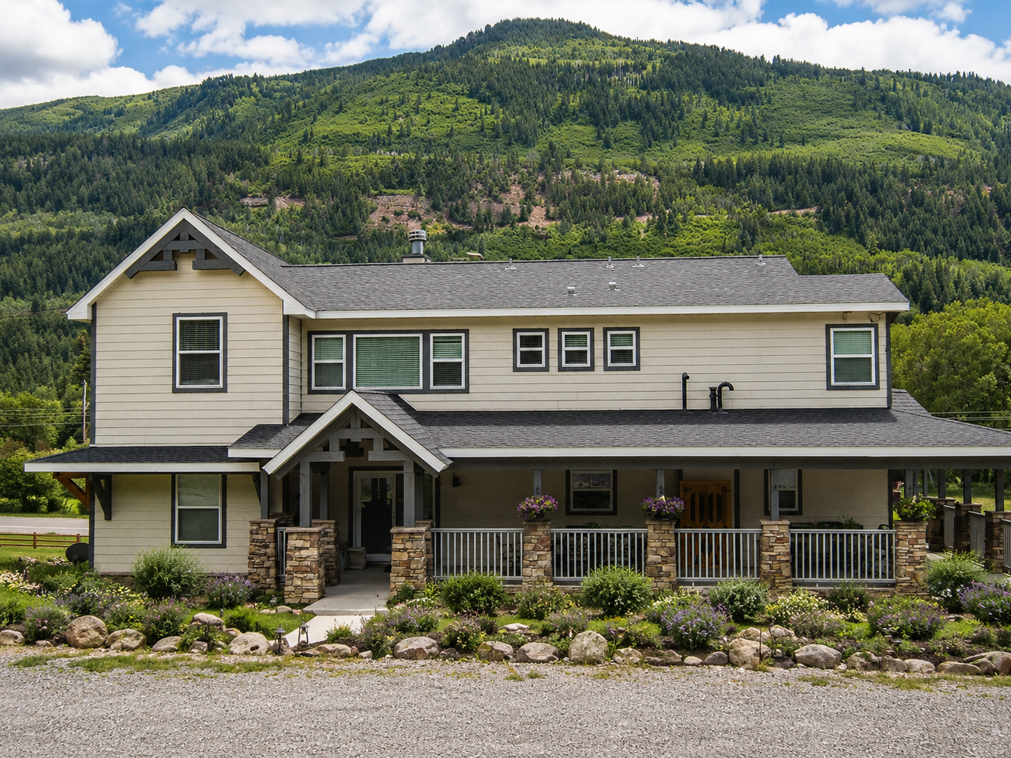 Two-story beige house with a porch, set before a forested mountain backdrop
