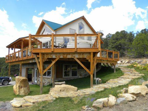 Wooden house with large wraparound deck on stilts beside a rocky path in a sunny rural setting