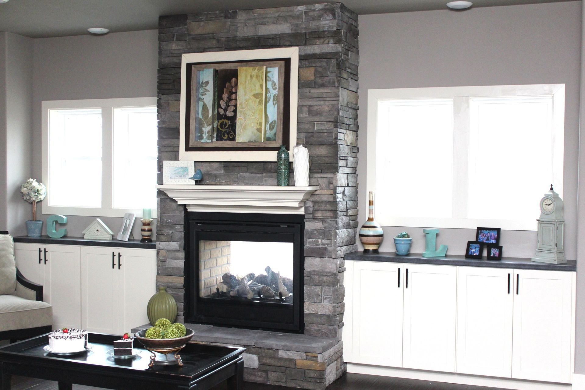 Modern living room with stone fireplace, framed art, white cabinets, and bright windows on both sides