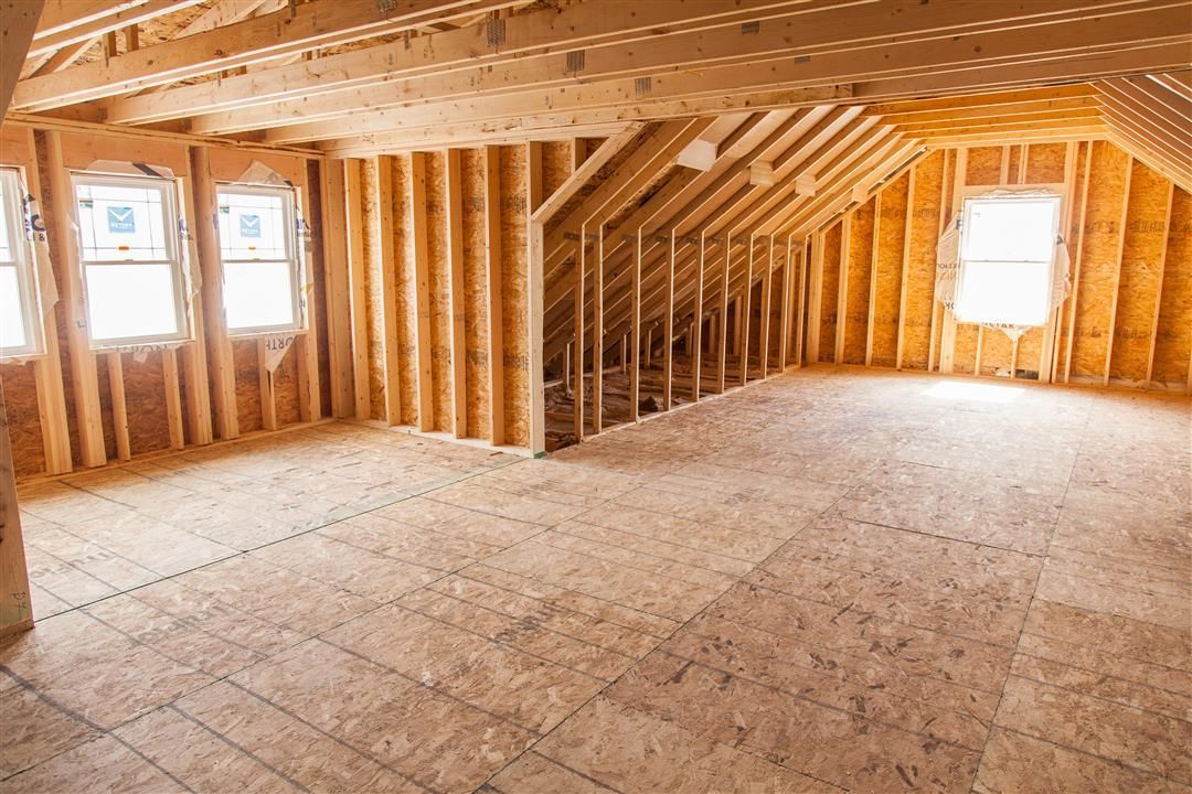 Unfinished attic interior with exposed wooden framing, plywood floor, and small windows letting in daylight