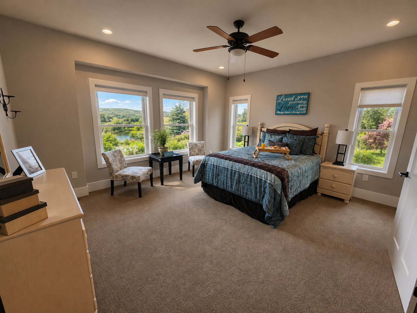 Bright bedroom with a bed, chair, dresser, ceiling fan, and large windows overlooking greenery