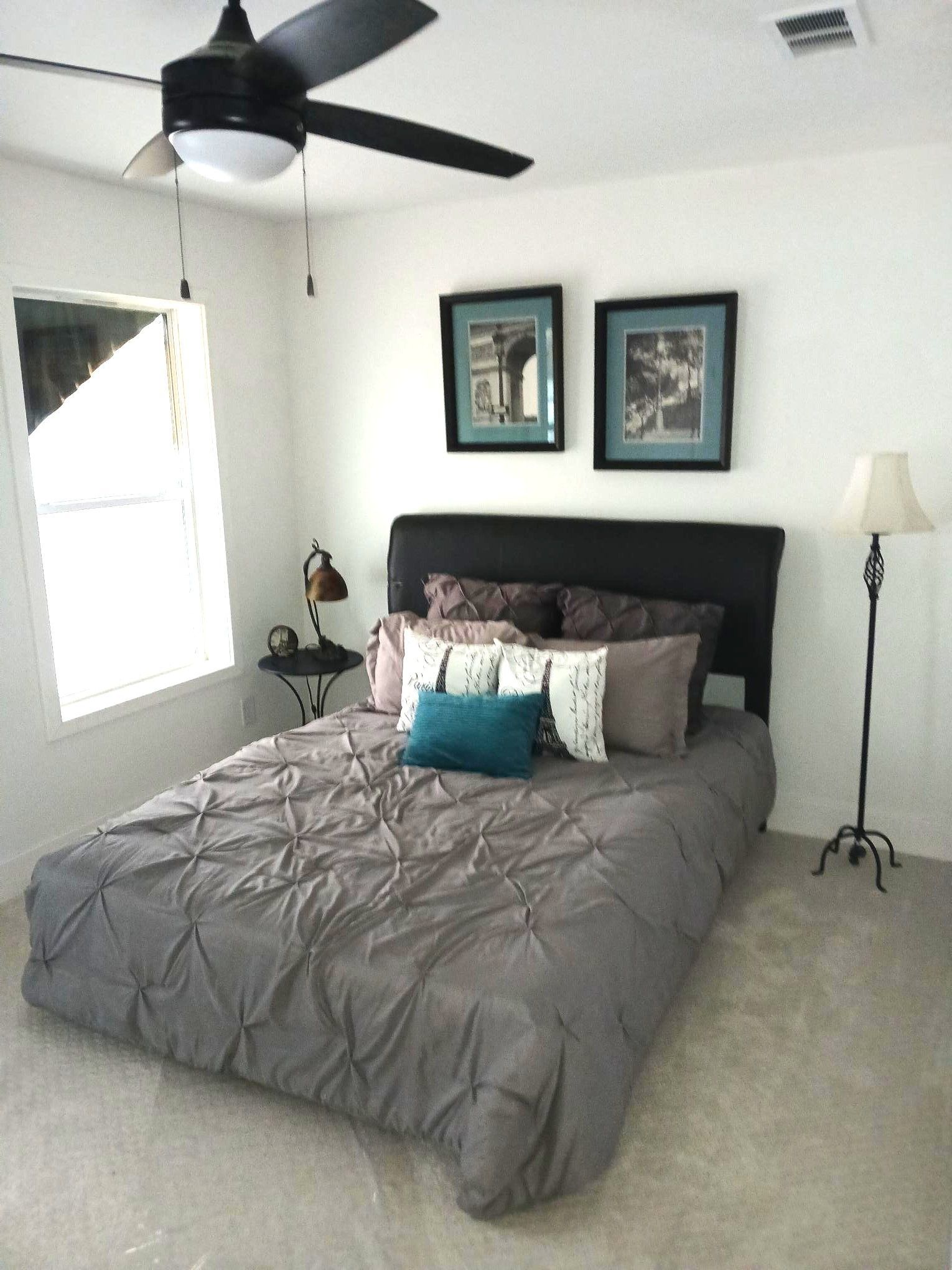 Bedroom with gray bedspread, black headboard, ceiling fan, and framed wall art.