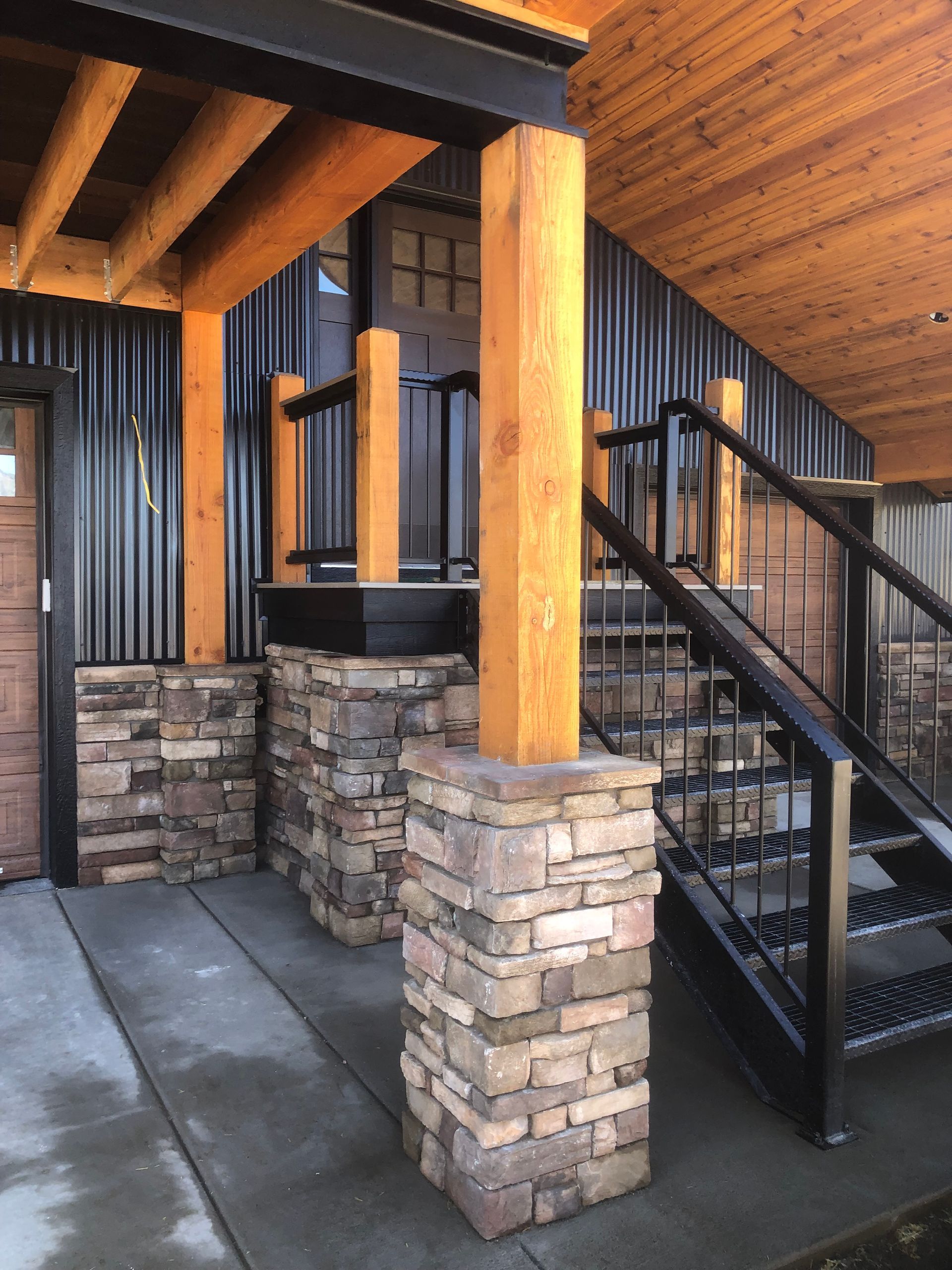 Covered outdoor porch with stone columns, black metal stairs, and wood ceiling and beams.