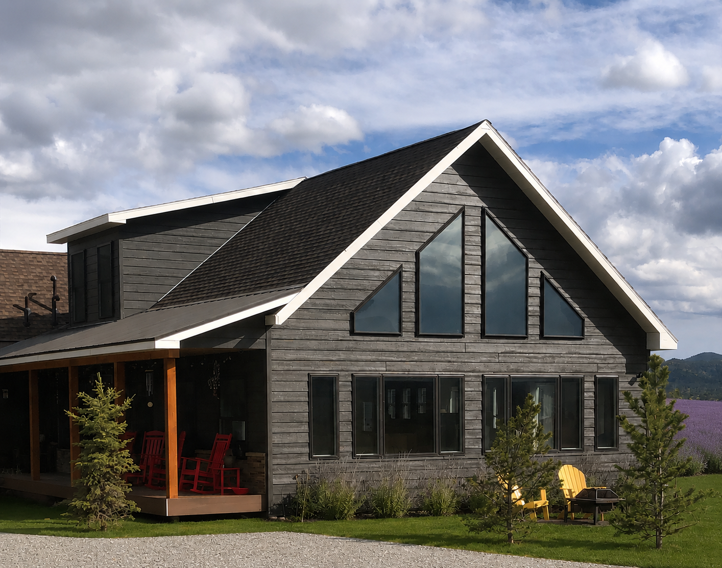 Modern gray house with a black roof, large windows, and a red porch on a gravel lot under a cloudy sky