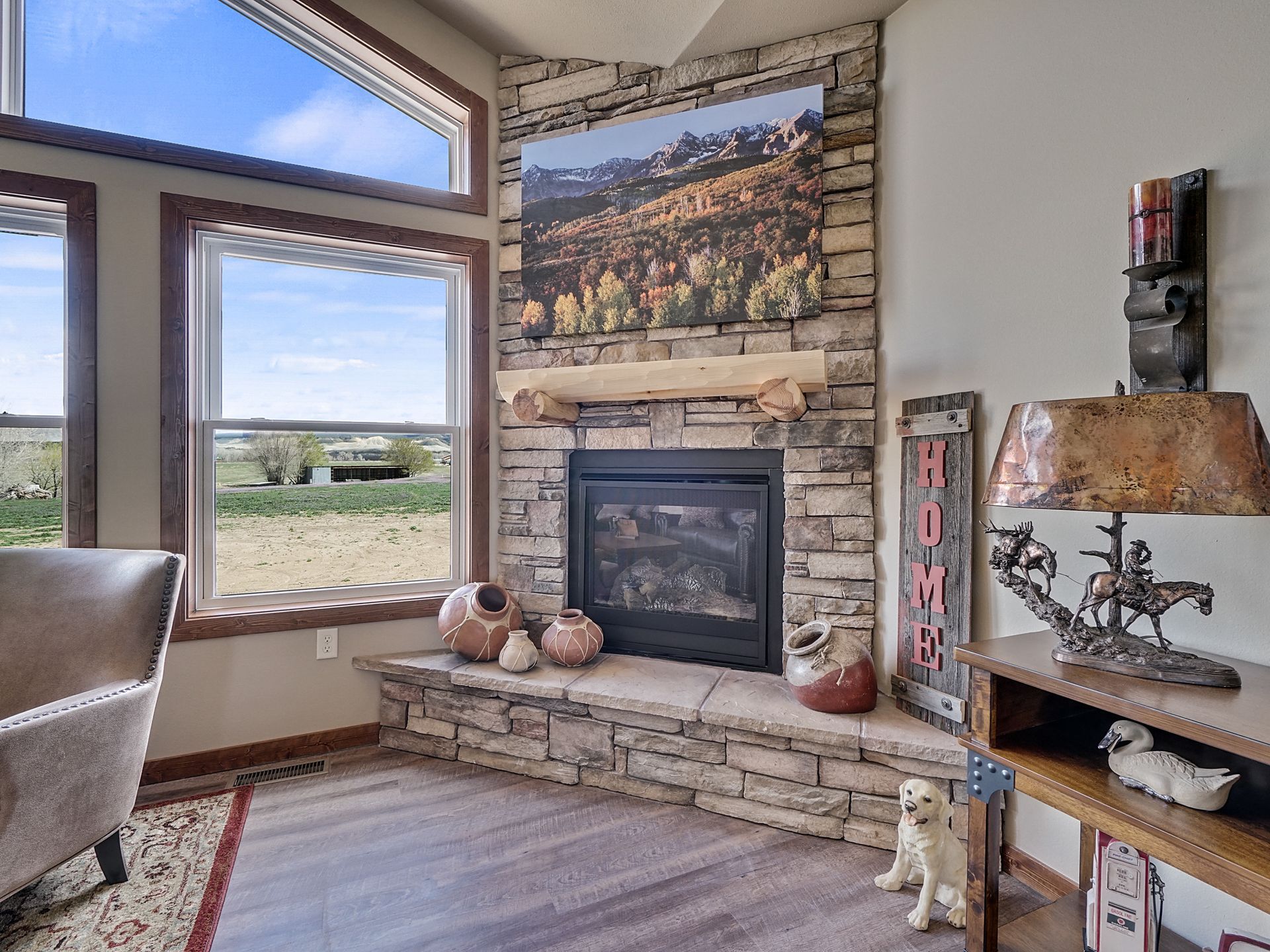 Living room with stone fireplace, large windows, landscape art, and rustic decor