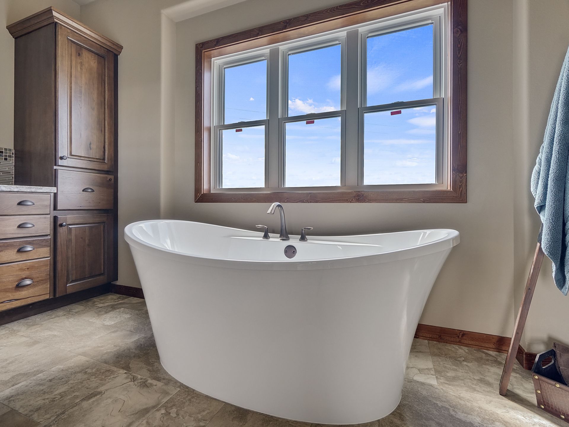 Freestanding white bathtub under a large window in a bright bathroom with wood cabinetry