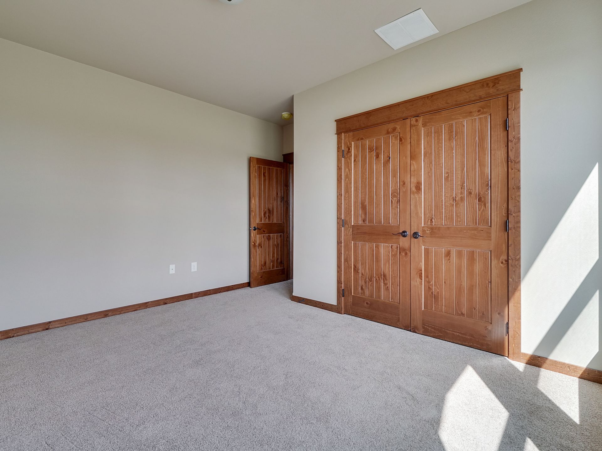 Empty beige bedroom with carpet, wooden double closet doors, and a sunlit wall