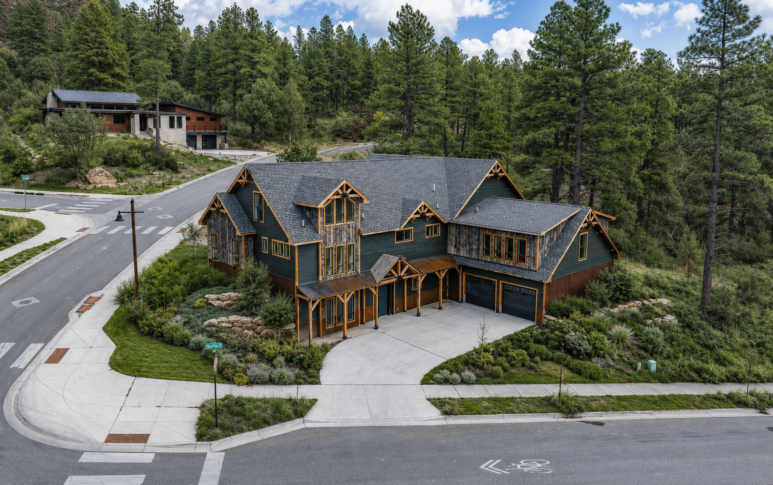 Large rustic mountain house with a dark roof, curved driveway, and wooded hillside surroundings