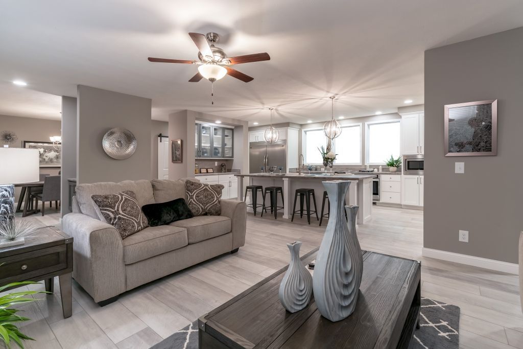 Bright open-plan living room with gray sofa, coffee table decor, ceiling fan, and kitchen island in the background