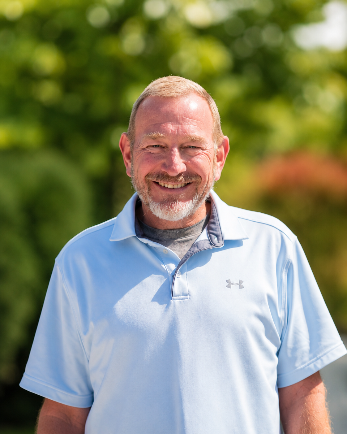 Smiling man in a light blue polo shirt standing outdoors with a blurred green background
