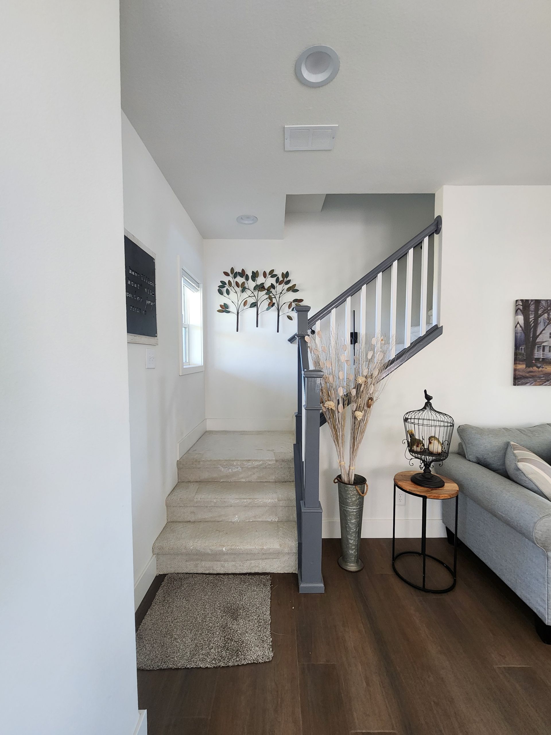 Bright entry hall with carpeted stairs, dark wood floor, gray sofa, and small side table with vase