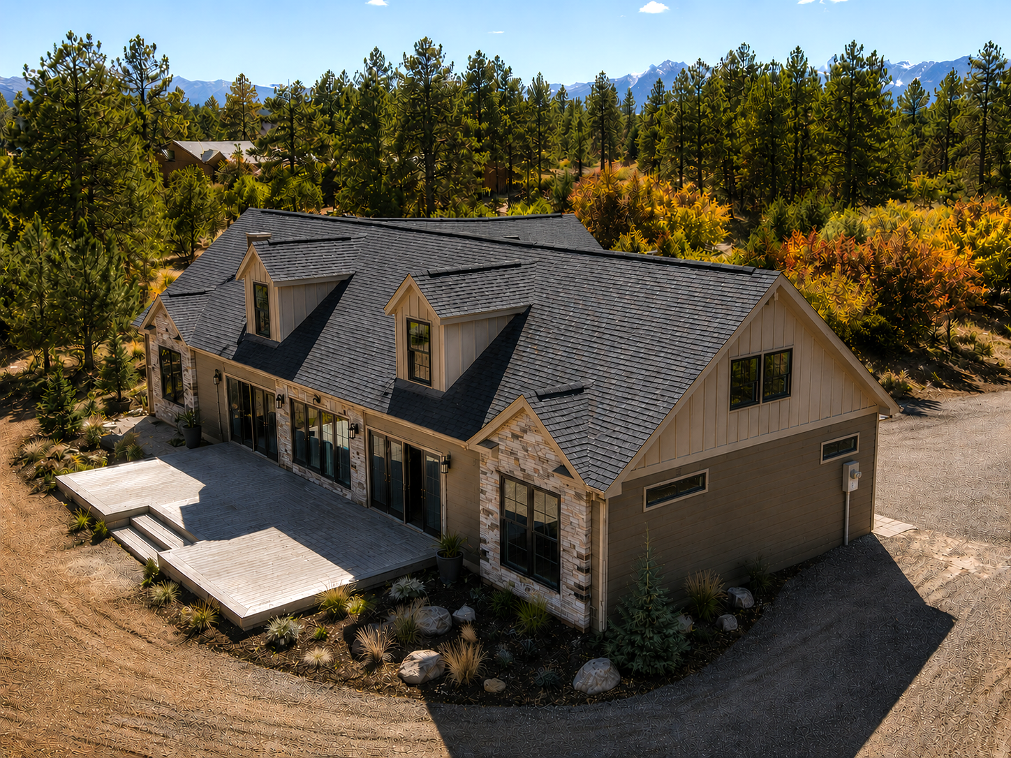 Large beige house with dark roof on a wooded lot, surrounded by autumn trees and gravel driveway