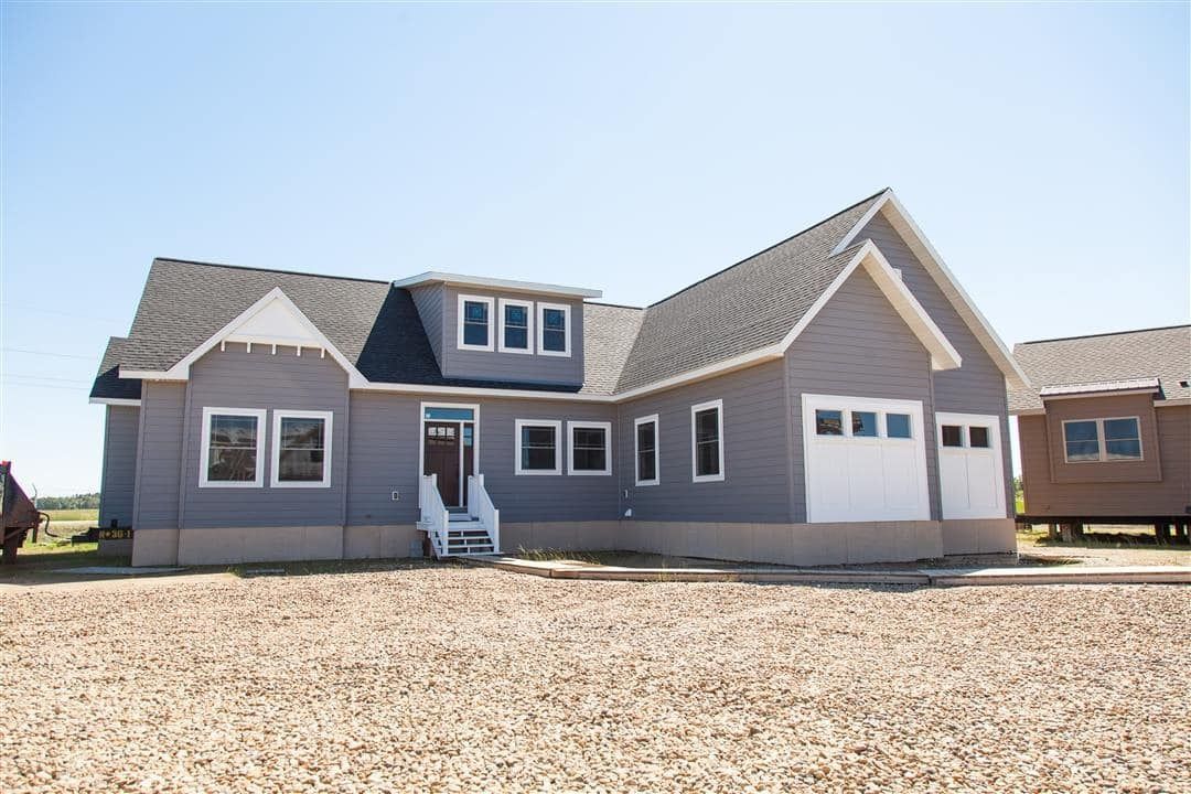 Gray house with white trim, front porch, and attached garage in a gravel lot under a clear sky
