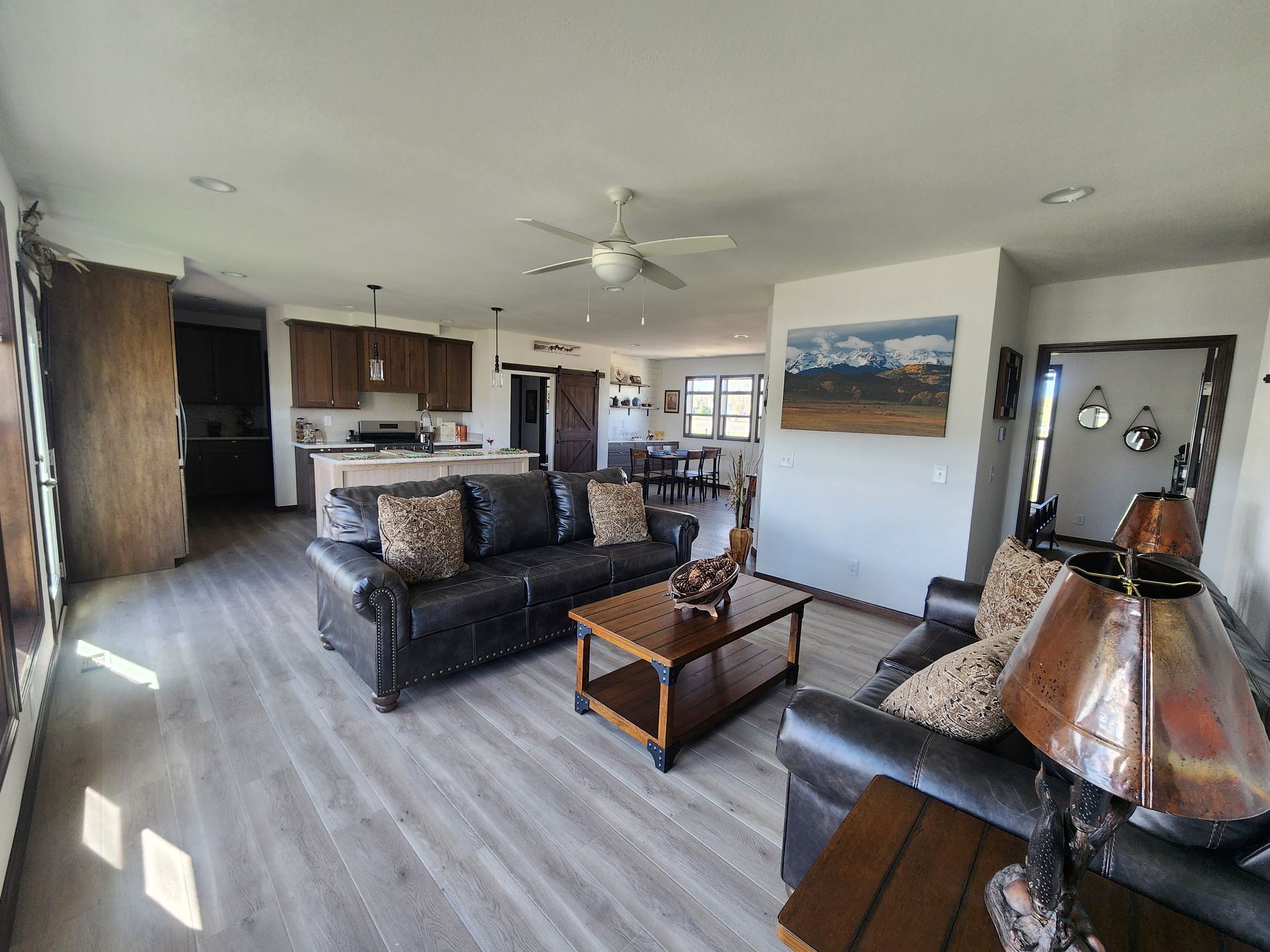 Open-plan living room and kitchen with dark sofas, coffee table, wood flooring, and ceiling fan