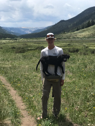 Person stands in a grassy mountain valley holding a black dog, with hills and forest in the background.