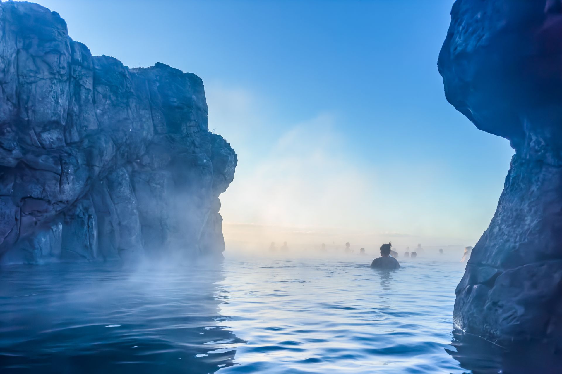 Person swimming in a misty, blue thermal pool, surrounded by rock formations, under a clear sky.
