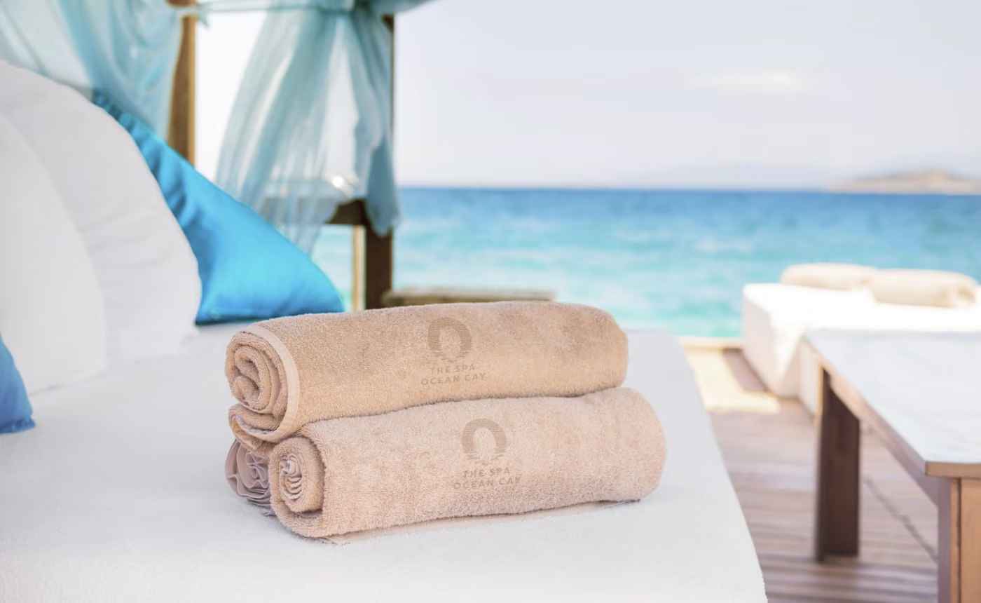 Rolled beige towels stacked on a bed with blue pillows and a view of the ocean.