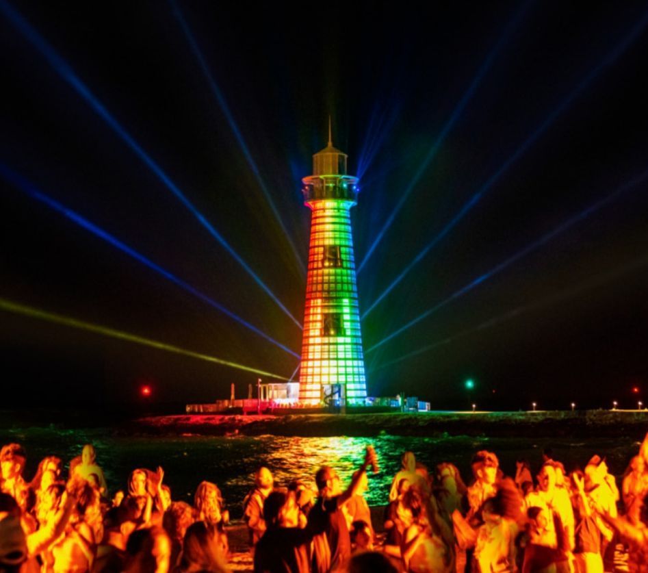 Lighthouse illuminated with rainbow lights at night, people in foreground.