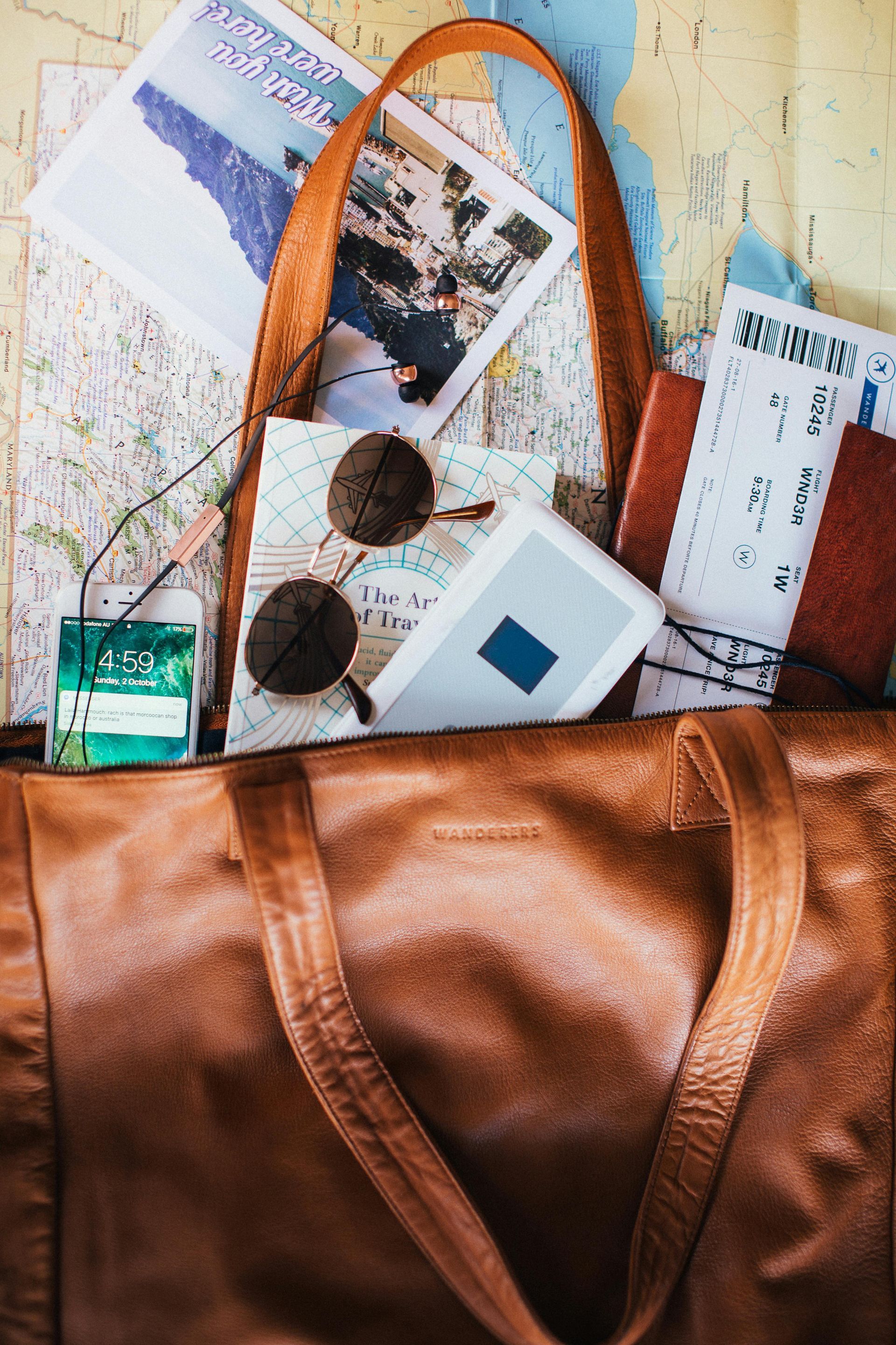 Brown leather travel bag overflowing with maps, sunglasses, documents, and hand sanitizer.