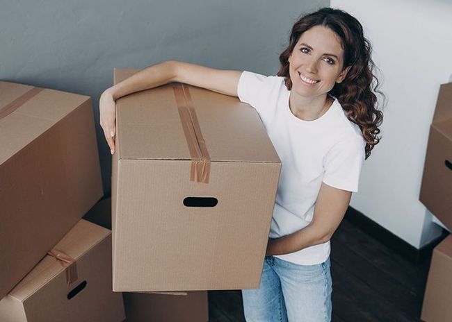 Smiling woman holding a cardboard moving box in a room with other packed boxes