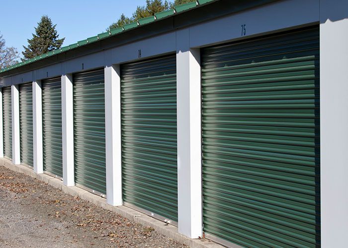 Row of green roll-up storage unit doors with white pillars along a gravel lot