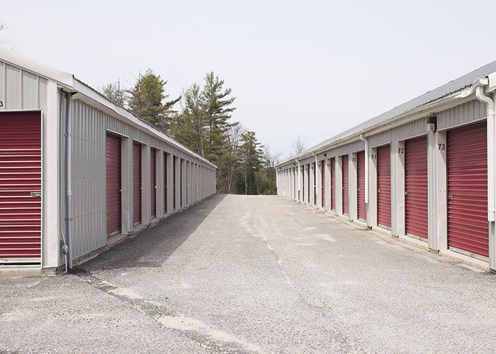 Row of red storage unit doors along a gravel lane, viewed between two self-storage buildings.