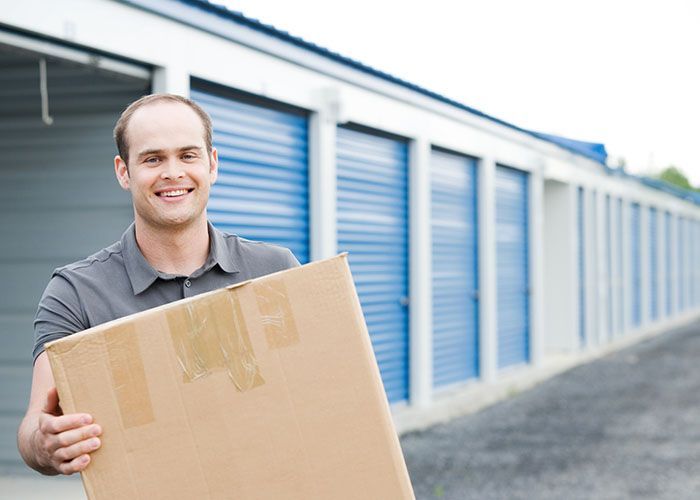 Man smiling while holding a large cardboard box outside blue storage units