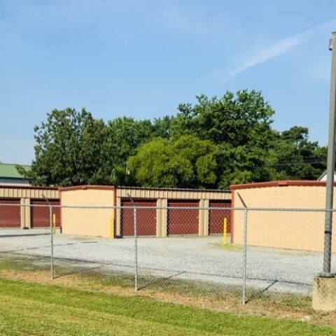 Chain-link fence in front of beige storage units and green trees under a clear blue sky