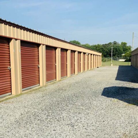 Gravel lane beside row of tan storage units with red roll-up doors under a clear blue sky