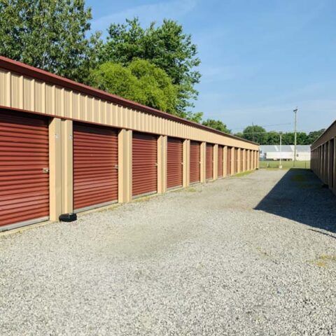 Row of red storage unit doors beside a gravel lane under a clear blue sky