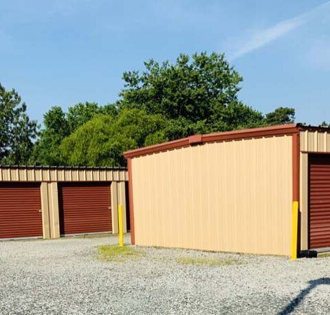 Row of storage unit garages with beige and red doors on a gravel lot under a clear blue sky