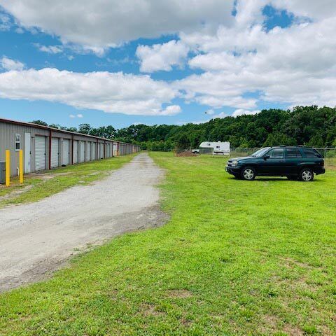 Gravel driveway beside storage units, with a black SUV parked on grass under a partly cloudy sky.