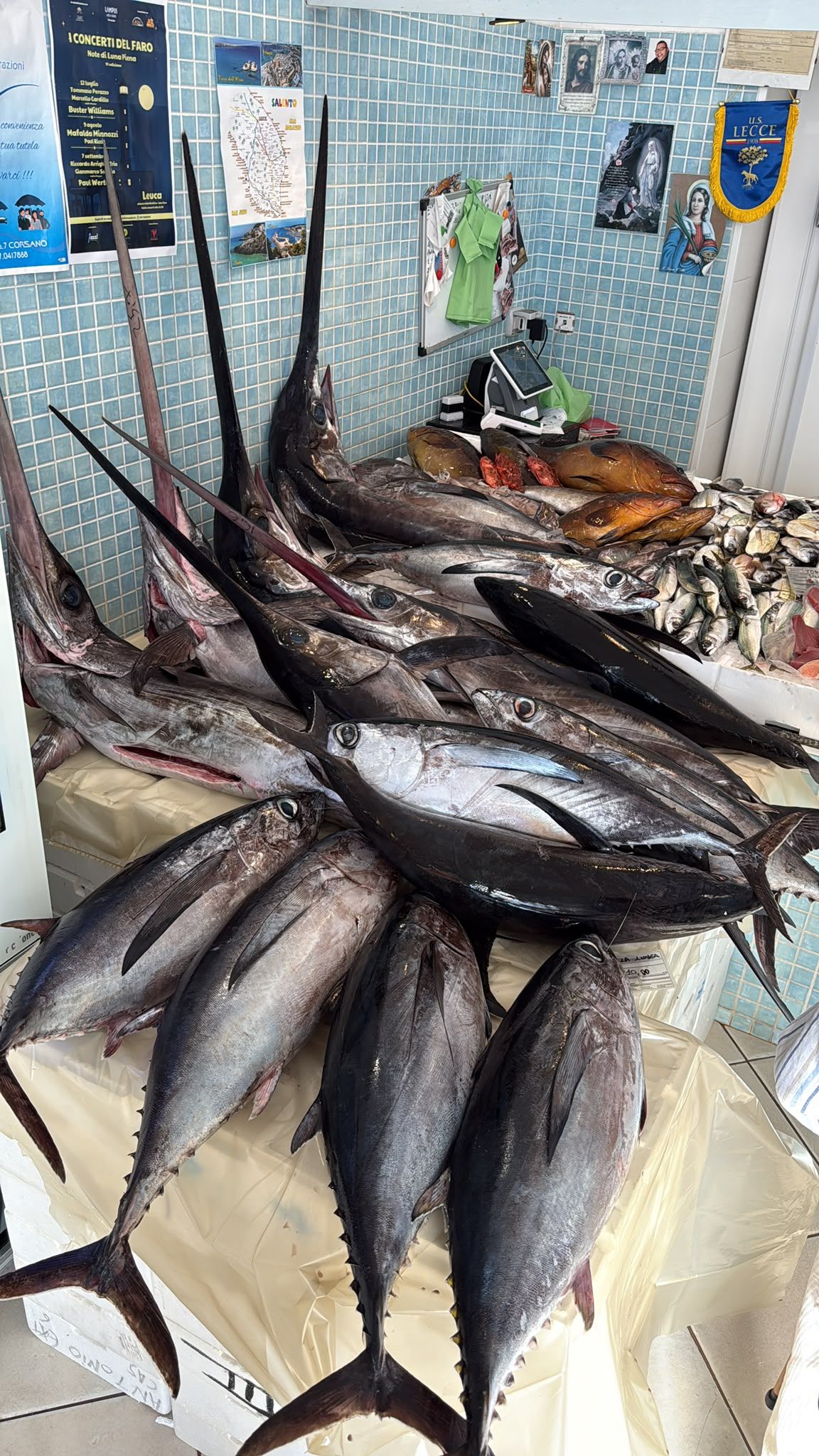 Pesce fresco esposto sul pavimento del mercato, con canne da pesca e un muro blu sullo sfondo.