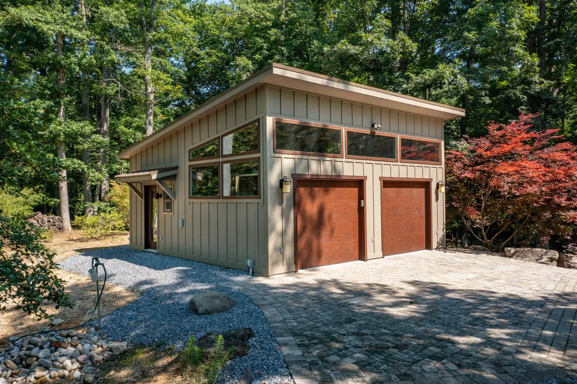 Beige garage with two brown doors, windows, and gravel driveway, surrounded by trees.