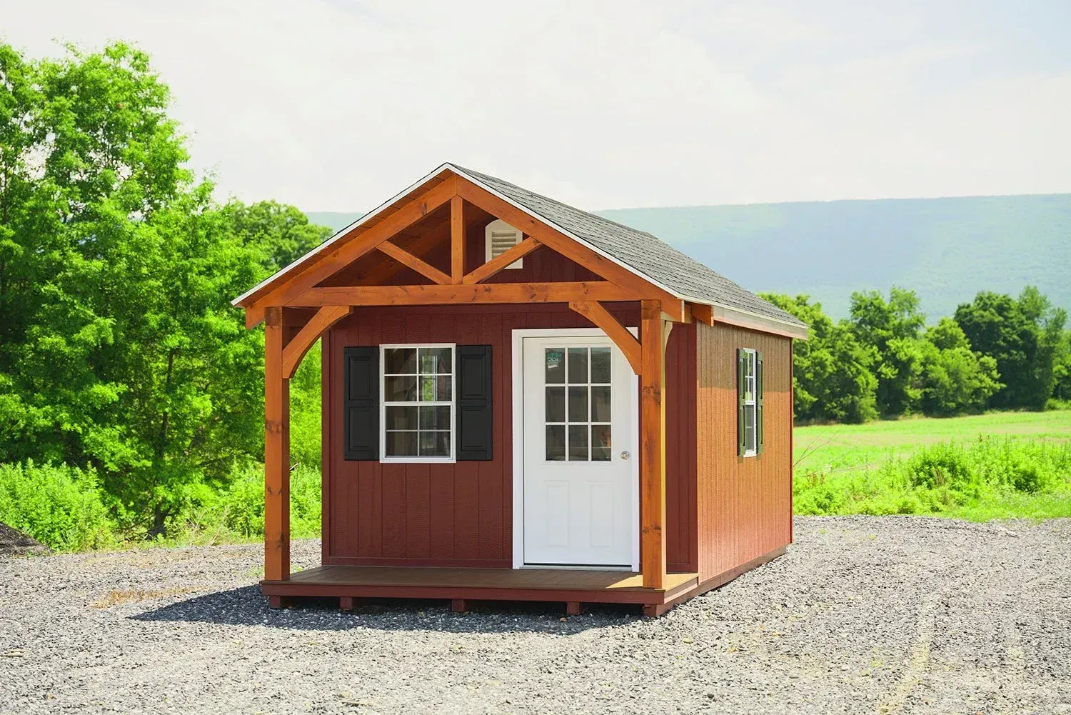 Small red cabin with porch, white door, and shutters. Set in a gravel area with trees and a mountain background.